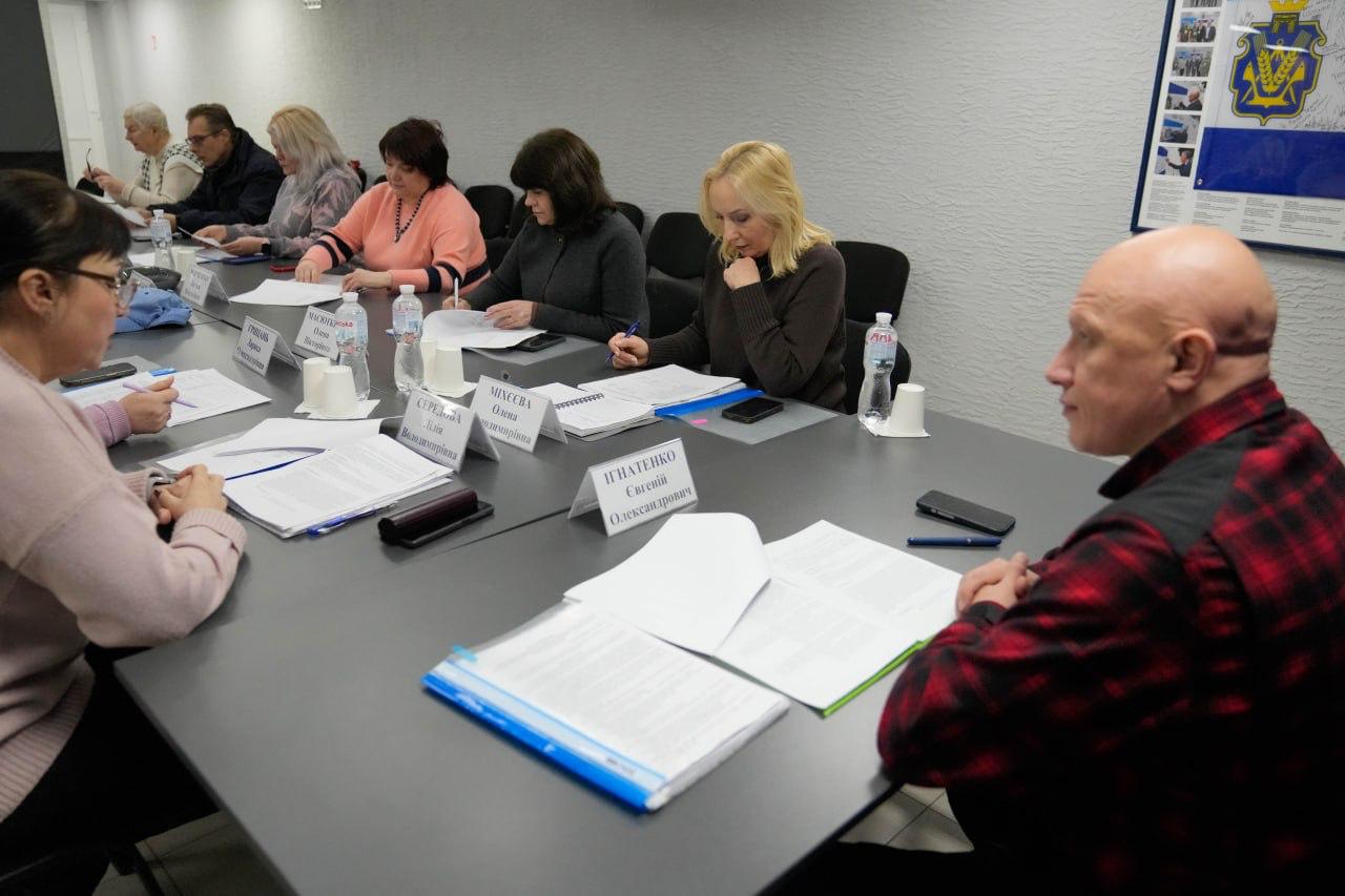 Учасники офіційного засідання сидять за робочим столом, вивчаючи документи в папках. Перед кожним присутнім стоять таблички з іменами та пляшки з водою. На передньому плані праворуч — чоловік у червоній клітчастій сорочці, який спостерігає за процесом обговорення.