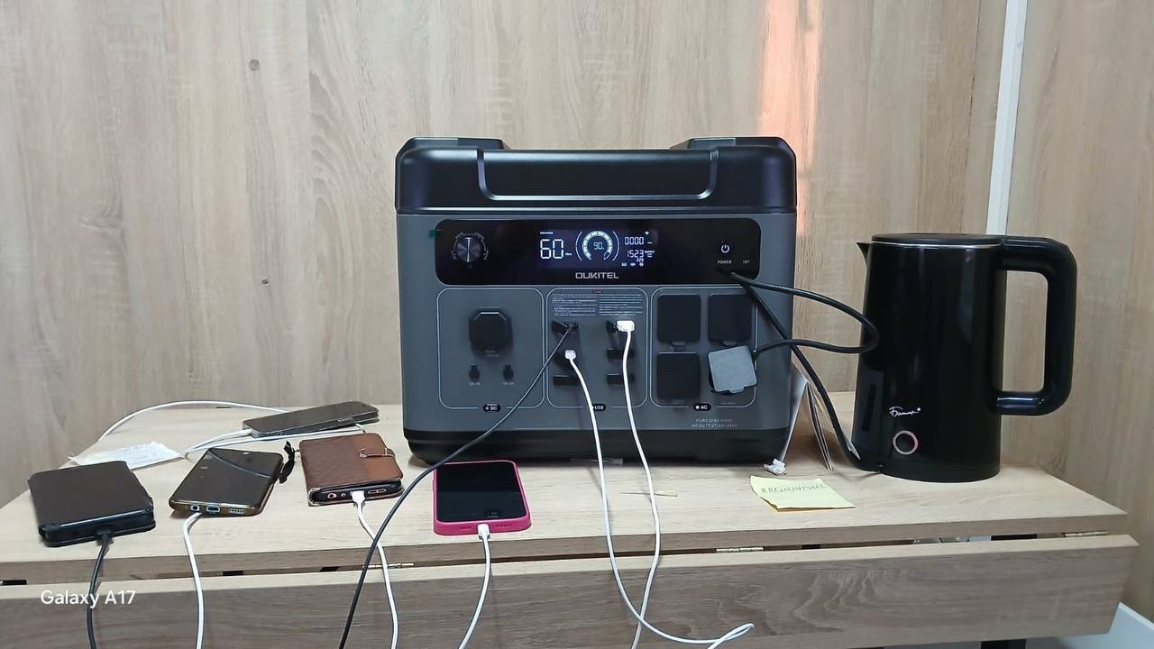 Зарядна станція Oukitel на дерев'яному столі. До пристрою підключено через USB-кабелі чотири смартфони, а через розетку змінного струму — електричний чайник чорного кольору. На цифровому дисплеї станції відображається рівень заряду 60%.