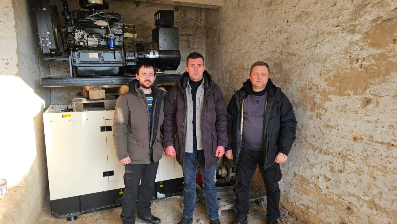 Троє чоловіків стоять у технічному приміщенні поруч із потужним дизельним генератором. Обладнання встановлене на спеціальній платформі. Стіни приміщення мають грубу текстуру, а підлога бетонна. Усі присутні одягнені в теплий верхній одяг.