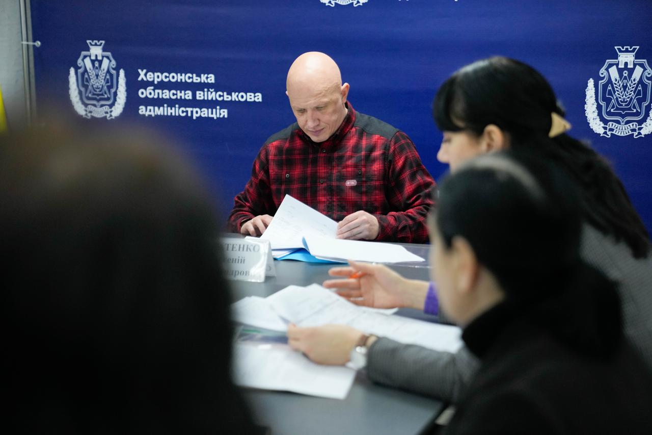 Великий план учасників наради в Херсонській обласній військовій адміністрації. У центрі — чоловік у червоній клітчастій сорочці, який переглядає документи за столом. На передньому плані видно силуети інших учасників обговорення, що працюють із паперами на фоні синього брендованого банера.