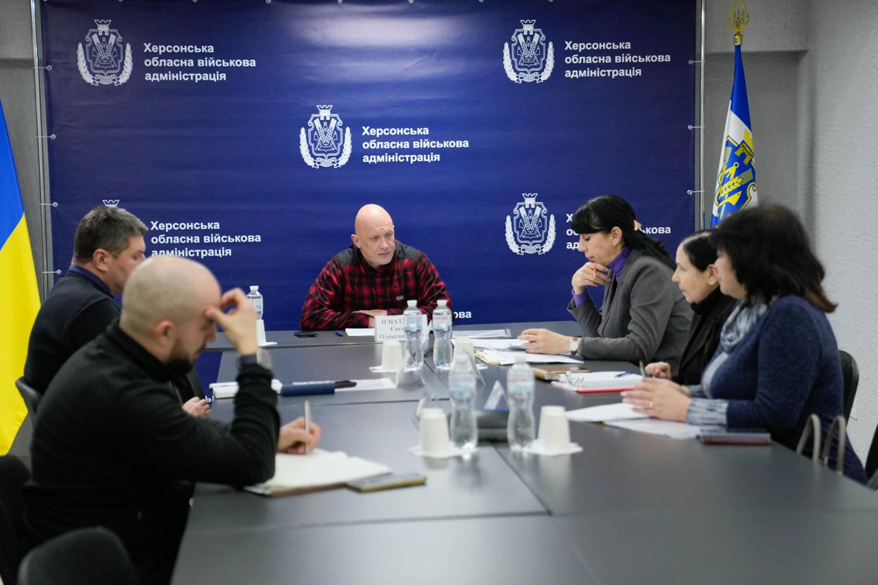 Робоча нарада в Херсонській обласній військовій адміністрації. Кілька людей сидять за довгим столом у конференц-залі на фоні синього банера з офіційними логотипами. У центрі — чоловік у червоній клітчастій сорочці, який веде обговорення, а інші учасники занотовують інформацію або слухають.