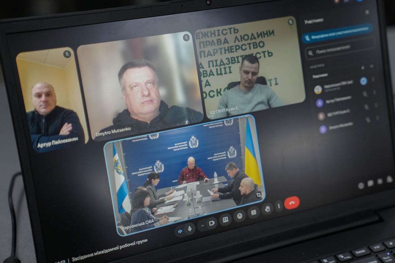 Екран ноутбука, на якому відображено вікно відеоконференції. У дзвінку беруть участь чотири вікна: три з окремими учасниками та одне із залу засідань Херсонської ОВА. Внизу екрана видно напис «Засідання міжвідомчої робочої групи».