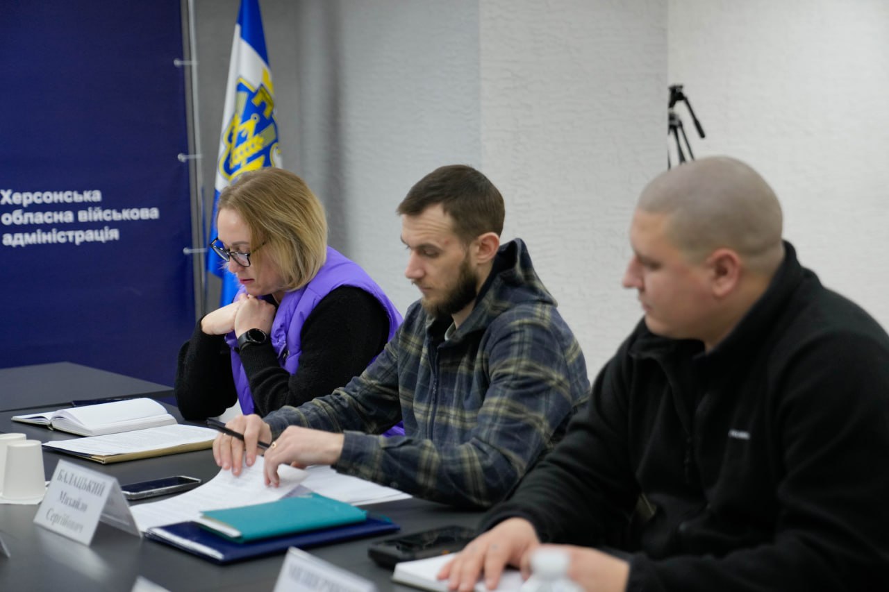 Троє учасників наради сидять за столом, вивчаючи документи та роблячи записи. На столі розміщені таблички з іменами та канцелярське приладдя.