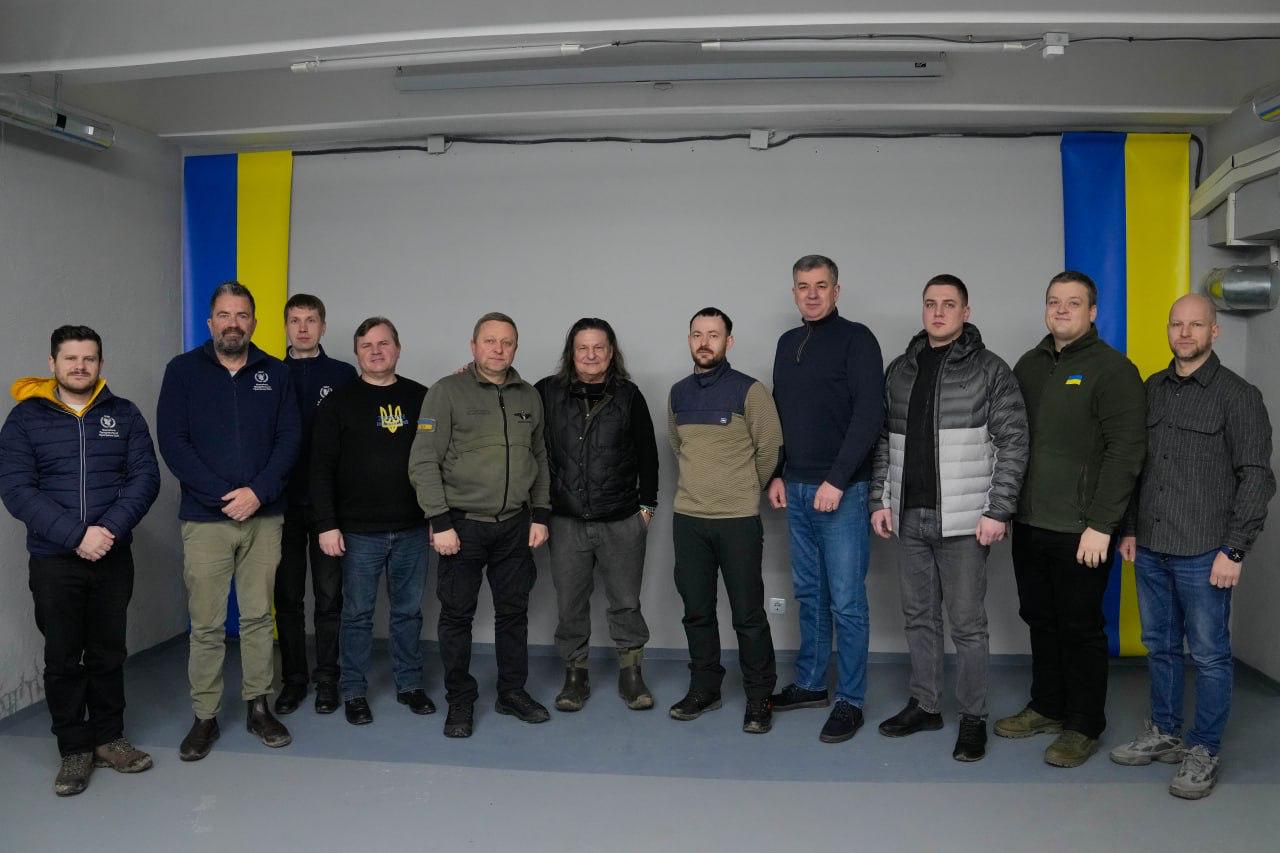 Група з одинадцяти чоловіків стоїть у ряд у світлому приміщенні на фоні сірої стіни. По обидва боки від них розвішані вертикальні синьо-жовті полотна, що нагадують прапор України. Більшість чоловіків одягнені у повсякденний або мілітарі одяг (худі, куртки, тактичні штани). Всі дивляться в об'єктив камери, атмосфера виглядає офіційною, але стриманою.