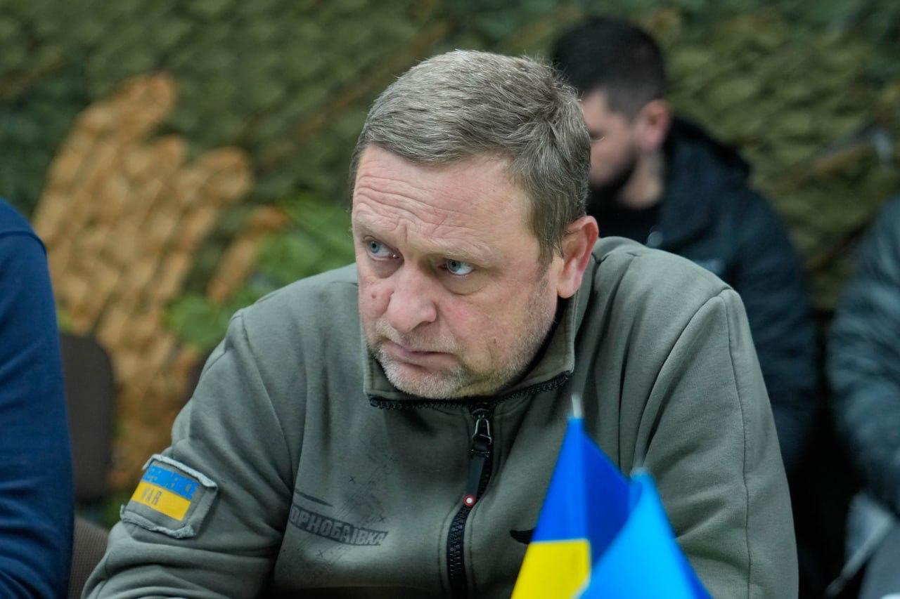 Портрет чоловіка у тактичній кофті кольору хакі з нашивкою у формі українського прапора на рукаві. Він має зосереджений та серйозний вираз обличчя, дивлячись трохи вбік. На фоні видно маскувальну сітку, а на передньому плані — частину невеликого синьо-жовтого прапорця.