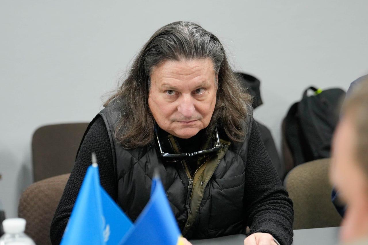 Портрет чоловіка з довгим волоссям, який сидить за столом. На ньому чорна стьобана жилетка та окуляри, що висять на комірі. Він дивиться прямо перед собою з уважним виразом обличчя. Перед ним на столі частково видно синій прапор ООН.