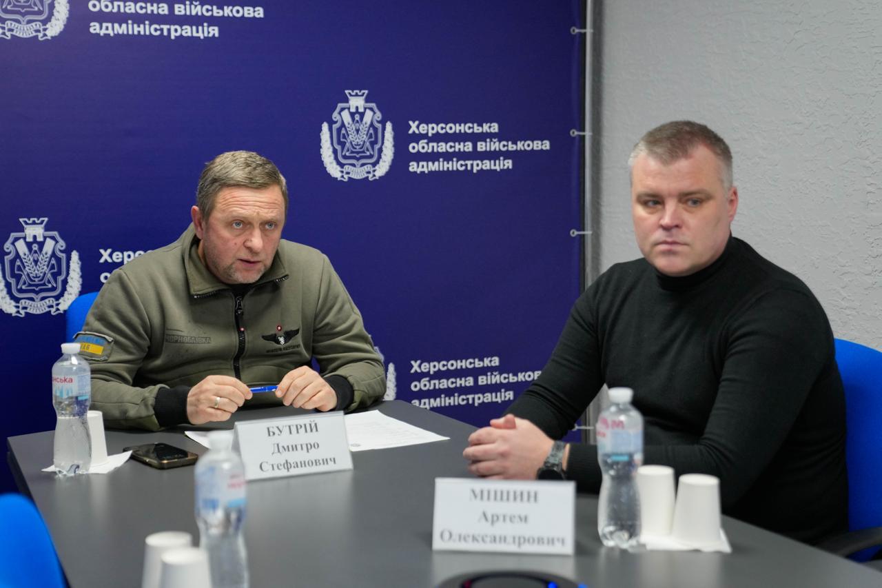 Двоє чоловіків за робочим столом на фоні синього банера з написом «Херсонська обласна військова адміністрація». Один у тактичному одязі кольору хакі з ручкою в руках, інший — у чорному светрі. На столі таблички з іменами та пляшки води.