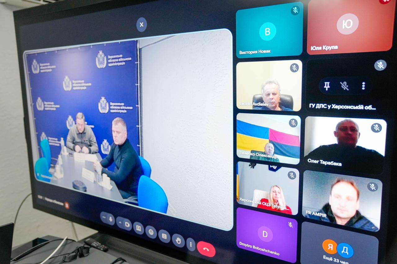 Екран монітора з онлайн-нарадою у форматі відеоконференції. У головному вікні — двоє чоловіків у залі із символікою Херсонської ОВА, у бічній панелі — плитки з іншими учасниками. Під кожним відео вказані імена або назви установ, більшість мікрофонів вимкнено.