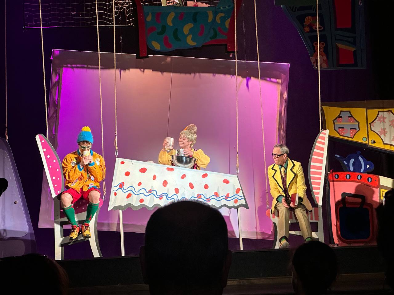 Сцена з театральної вистави у яскравих декораціях. Троє акторів у кольорових костюмах сидять за столом, що ніби підвішений на мотузках. На столі — біла скатертина у червоний горошок. Декорації навколо нагадують дитячі малюнки або сторінки книги.