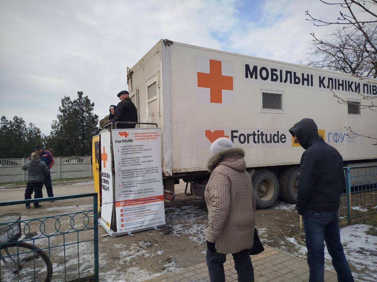 Біла вантажівка мобільної медичної клініки з написом «Мобільні клініки» та логотипами організацій, серед яких «Fortitude». Біля входу до фургона зібралися люди, на передньому плані видно інформаційний стенд із правилами надання гуманітарної допомоги.