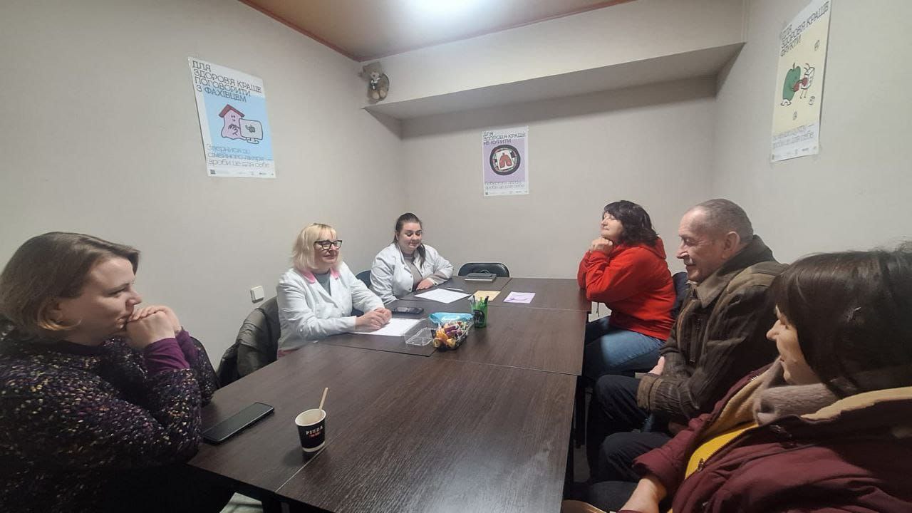 Група людей сидить за великим столом у кабінеті під час консультації або зустрічі. Двоє медичних працівників у білих халатах спілкуються з відвідувачами. На стінах розвішані плакати на медичну тематику з порадами щодо здорового способу життя.