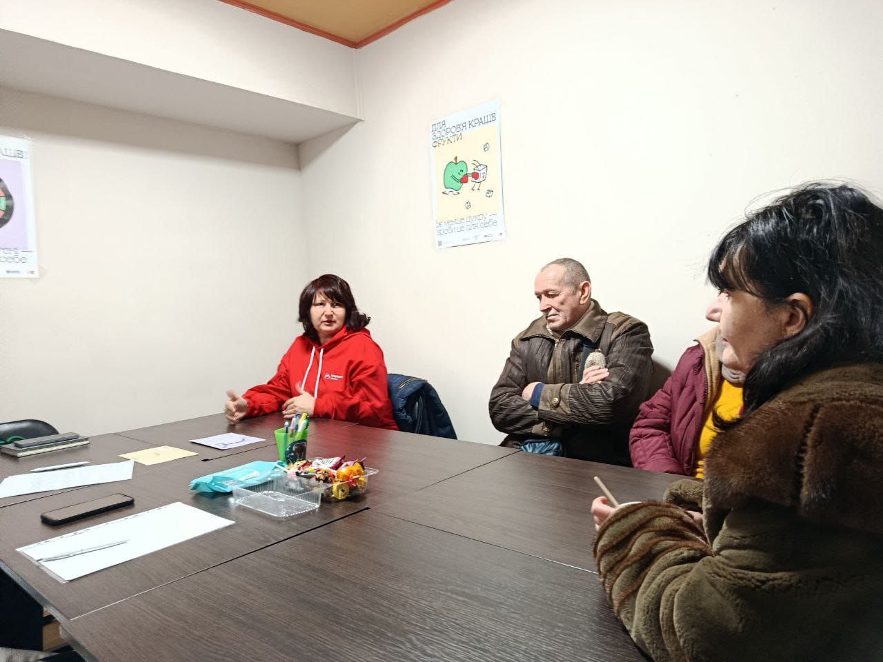 Учасники зустрічі сидять за столом під час обговорення. На стіні над ними розміщено плакат із порадами щодо здорового харчування, на якому зображено яблуко та напис «Для здоров'я краще фрукти». На столі лежать робочі матеріали, мобільний телефон та ємність із солодощами.