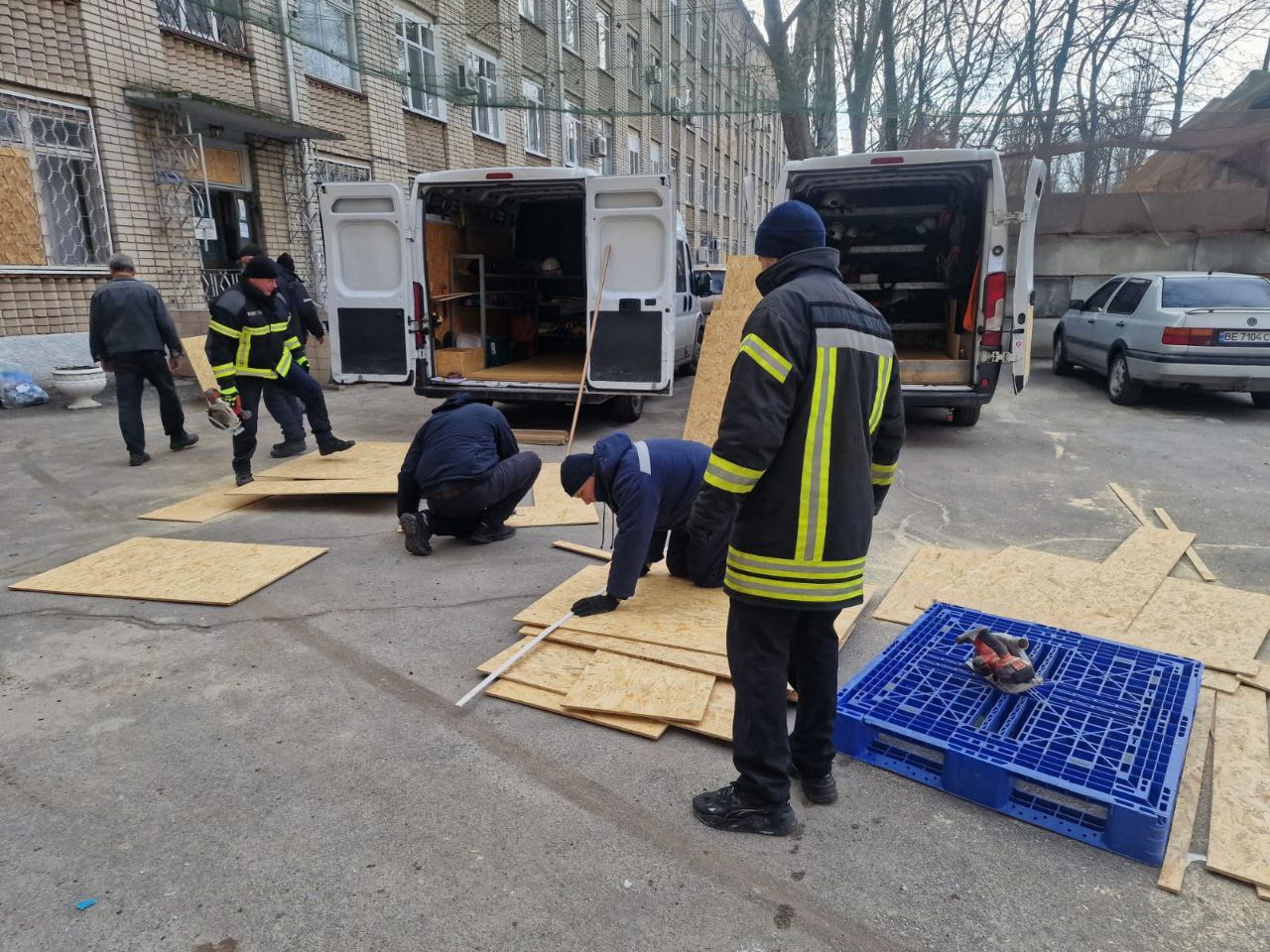 На подвір'ї біля пошкодженої цегляної будівлі працює група людей у спеціальному одязі. Двоє чоловіків у формі з яскравими світловідбивними смугами та інші працівники готують листи фанери, вимірюючи та розрізаючи їх. Поруч стоять два білі мікроавтобуси з відчиненими задніми дверима, завантажені інструментами та матеріалами для ремонтних робіт.