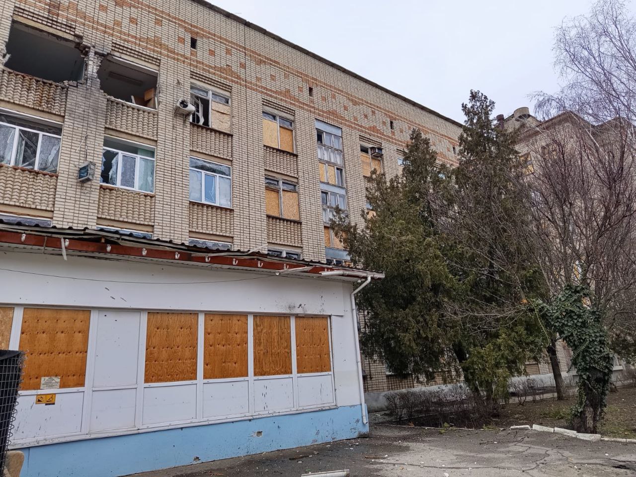 Цегельна багатоповерхова будівля з наслідками значних руйнувань. На фасаді видно вибиті шибки, а багато віконних прорізів забито фанерою або дерев’яними щитами. У верхньому лівому куті помітні пошкодження стін та міжповерхового перекриття. Поруч із будівлею ростуть дерева, а прилегла територія вкрита дрібним будівельним сміттям.