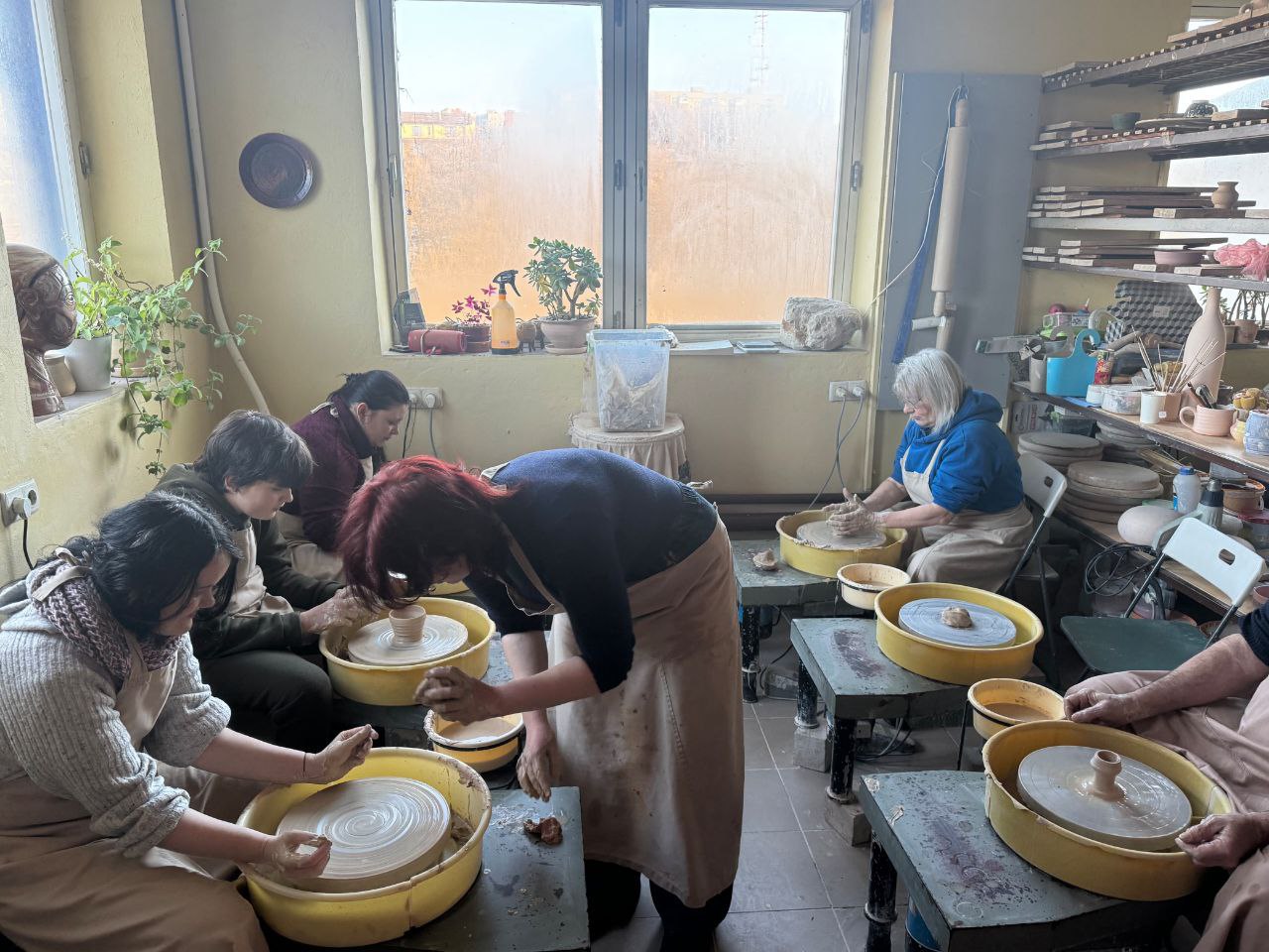 Група людей у захисних фартухах працює за гончарними колами в майстерні. Вони зосереджено формують вироби з глини. Приміщення заповнене стелажами з керамікою, рослинами на підвіконні та природним світлом із великого вікна.