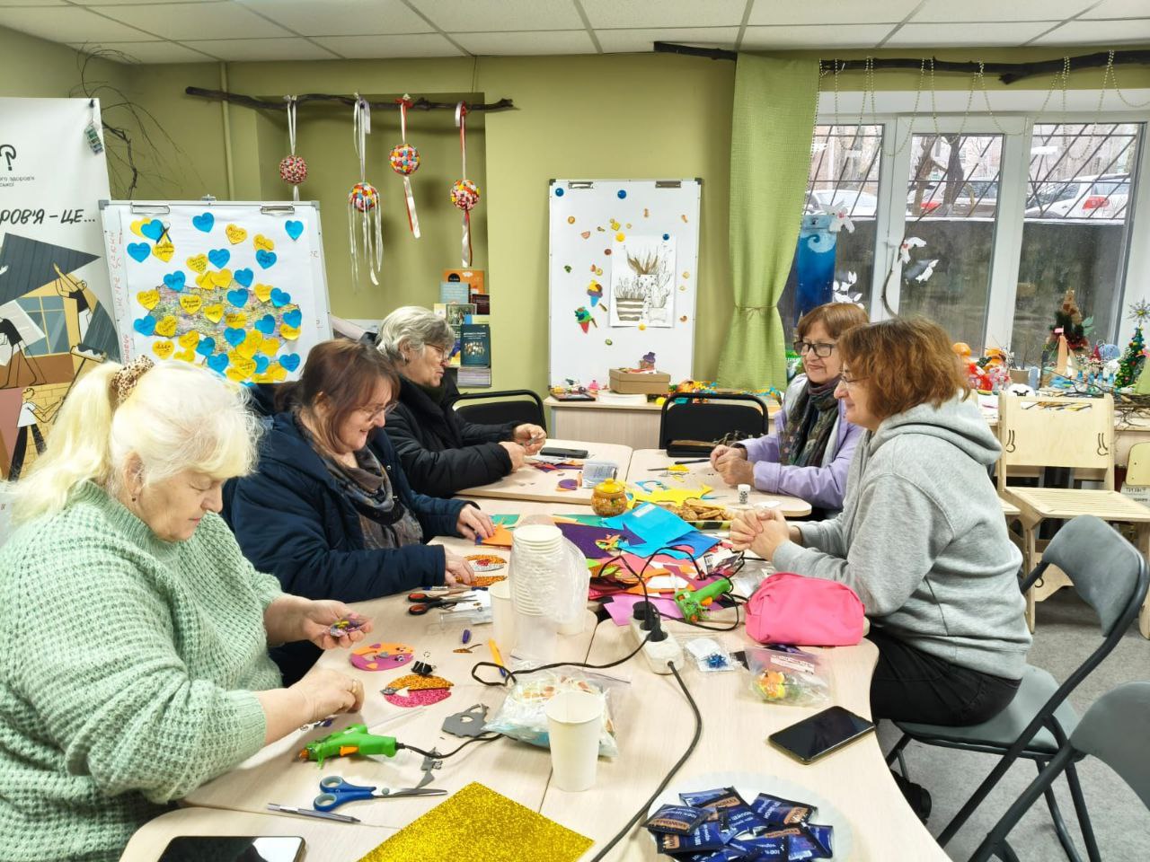 Група жінок старшого віку займається рукоділлям за спільним столом у світлому приміщенні. На столі розкладені кольоровий папір, інструменти та готові вироби. На фоні видно дошку з наліпками у формі серця та святковий декор.