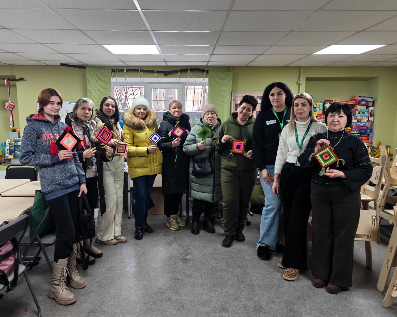 Група жінок різного віку стоїть у світлій кімнаті, демонструючи власноруч виготовлені обереги з кольорових ниток у формі ромбів. Вони одягнені в повсякденний зимовий та демісезонний одяг. На фоні видно робочі столи, стелажі з іграми та вікно з зеленими шторами.