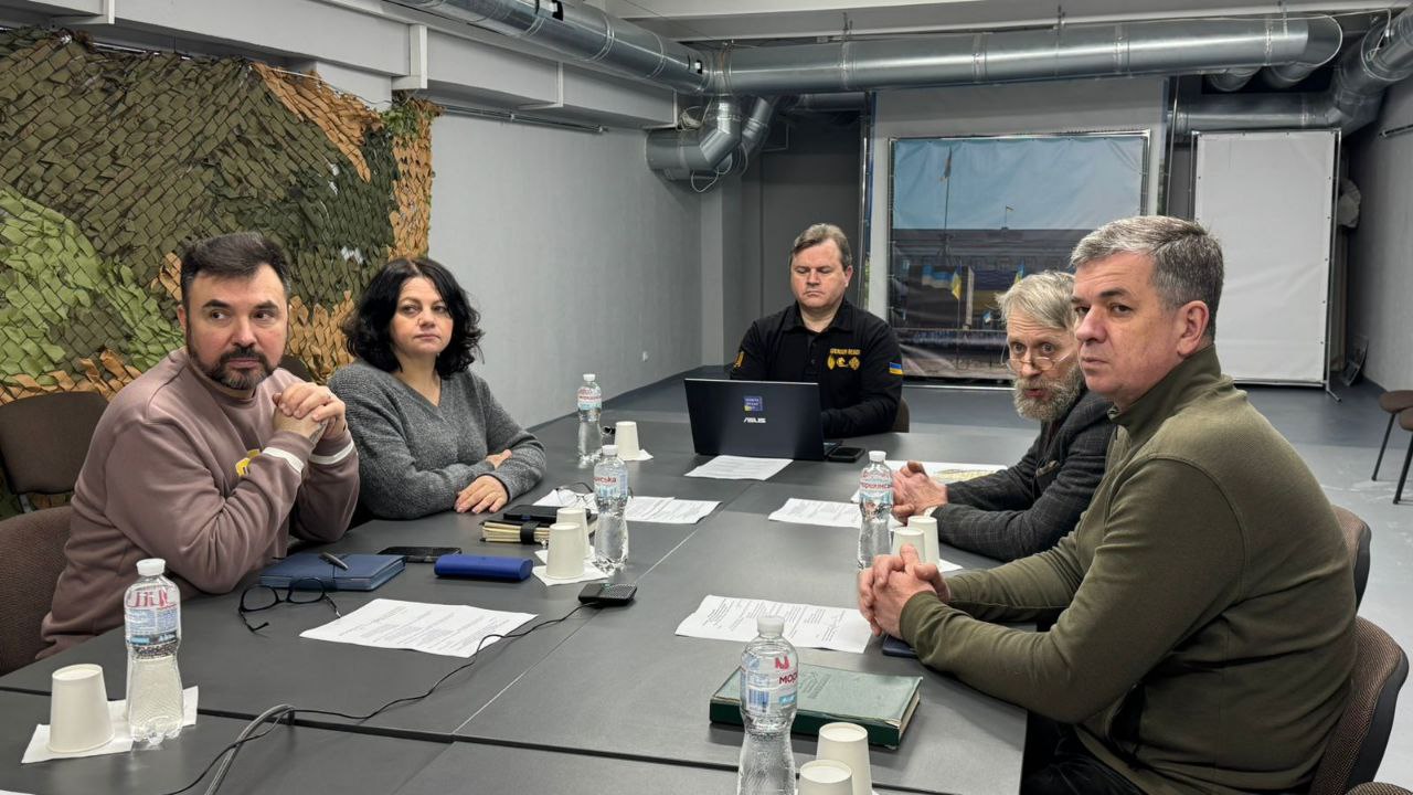 П'ятеро людей сидять за довгим столом під час робочої наради у технічному приміщенні з видимими комунікаціями на стелі. На столі розкладено папери, ноутбук та пляшки з водою, а на стіні ліворуч закріплена захисна маскувальна сітка.