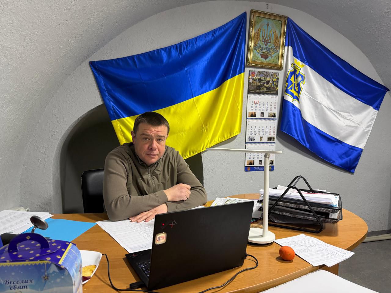 Чоловік у військовій формі сидить за робочим столом із ноутбуком у приміщенні зі склепінчастою стелею. На стіні за ним розміщені прапор України, прапор Херсонської області, державний герб та календар.