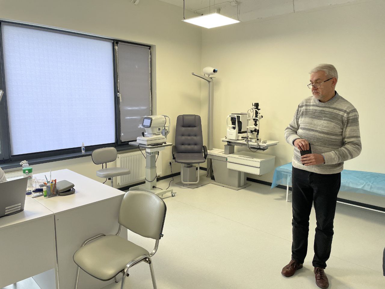 Чоловік у світлому светрі стоїть у сучасному офтальмологічному кабінеті. У приміщенні встановлено професійне обладнання для перевірки зору, спеціальне крісло для пацієнта, робочий стіл та кушетка.