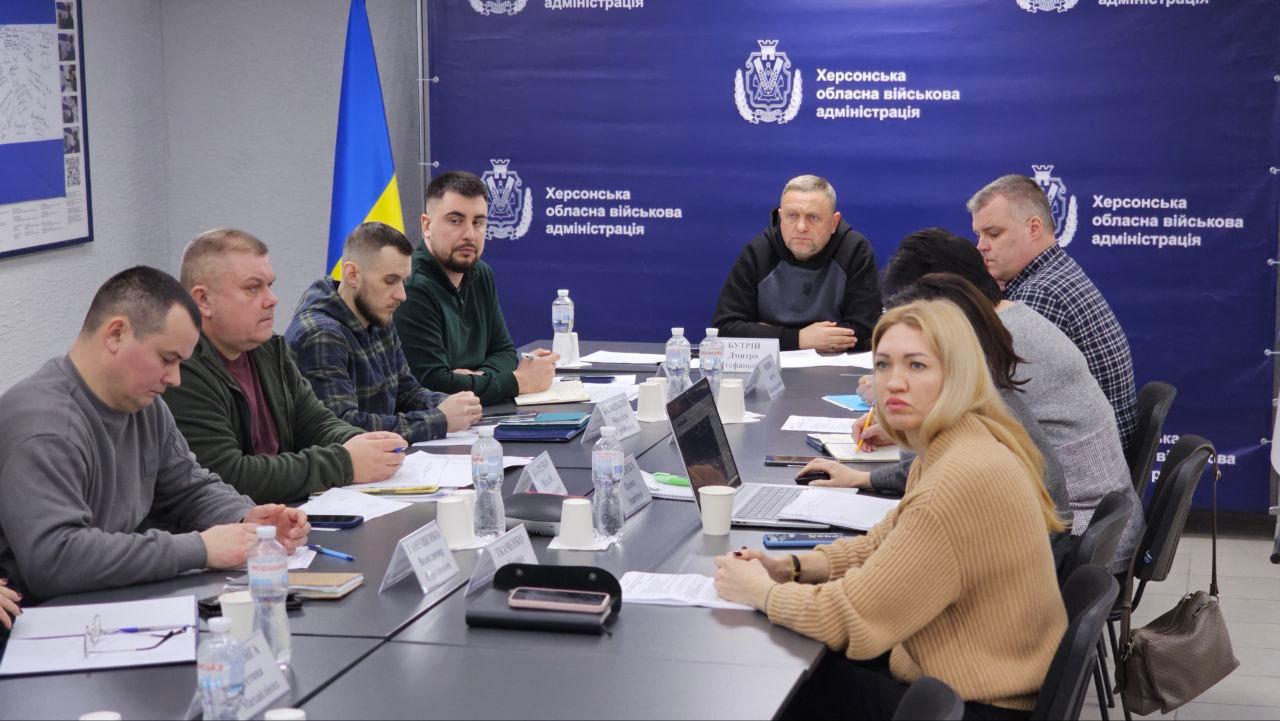 Група людей сидить за великим столом під час робочої наради у приміщенні Херсонської обласної військової адміністрації. На фоні розташований синій банер із офіційними логотипами установи та державний прапор України.