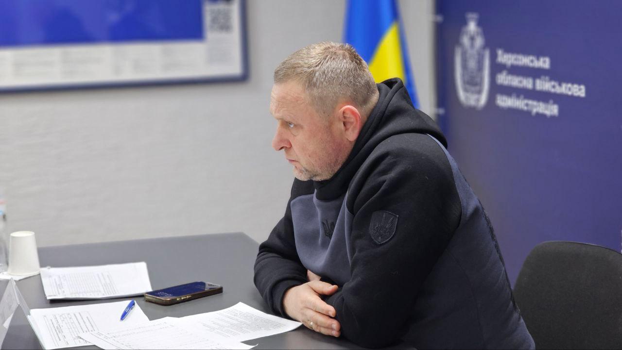 Чоловік у темному спортивному одязі сидить за столом у профілі, зосереджено дивлячись перед собою. Перед ним лежать робочі документи та ручка. На задньому плані видно частину синього банера з офіційною символікою та державний прапор України.