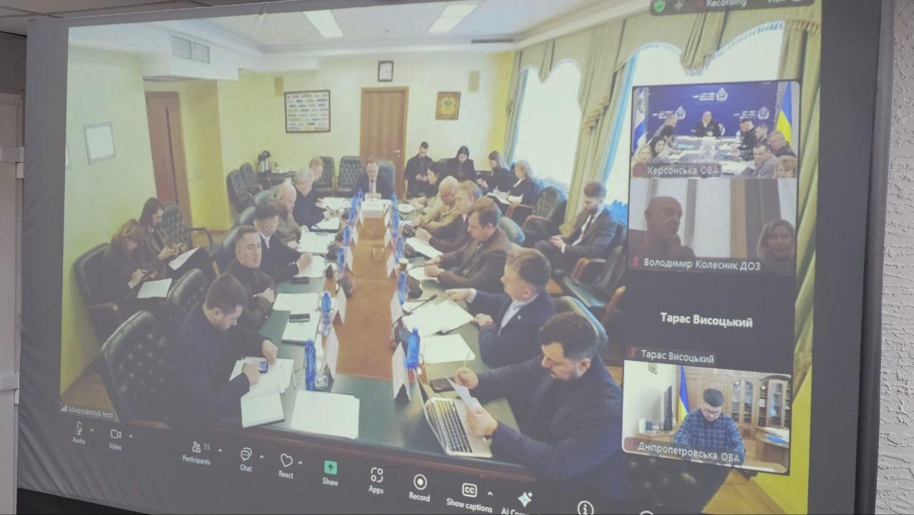 Фото екрана під час селекторної наради у форматі відеоконференції. На великому моніторі відображається зал засідань із учасниками за столом, а в боковій панелі — окремі вікна з іншими підключеними представниками обласних адміністрацій та відомств.