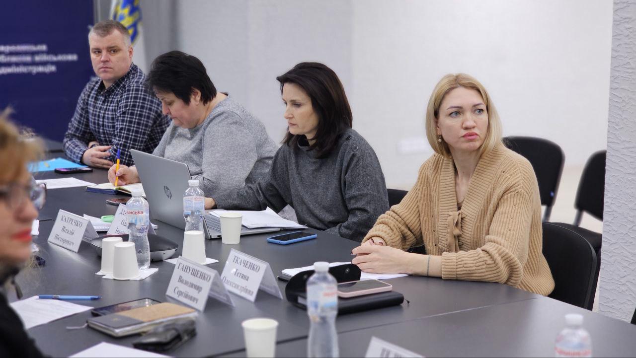 Учасники офіційної зустрічі сидять за робочим столом, уважно слухаючи виступаючого. На столі розміщені ноутбуки, пляшки з водою та таблички з іменами, а на фоні помітно частину офіційного банера установи.