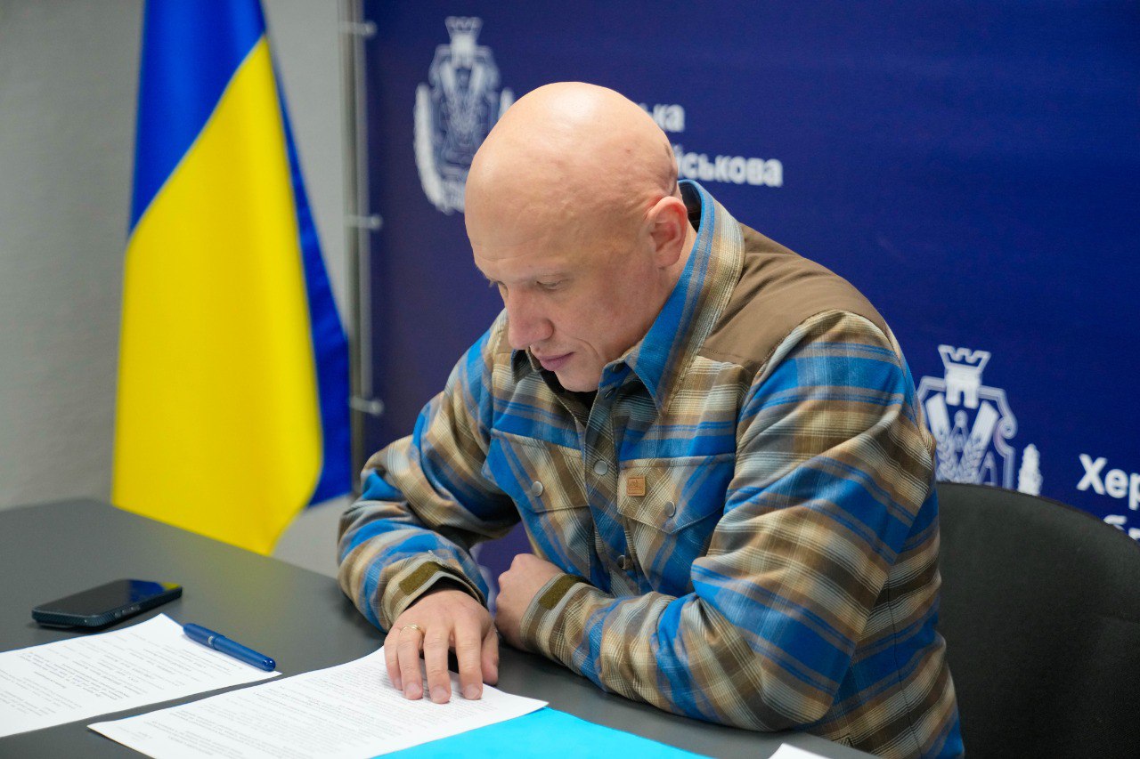 Чоловік у синьо-коричневій сорочці в клітинку сидить за робочим столом і зосереджено читає документи. Позаду нього розташований синій банер з логотипом Херсонської обласної військової адміністрації та прапор України.