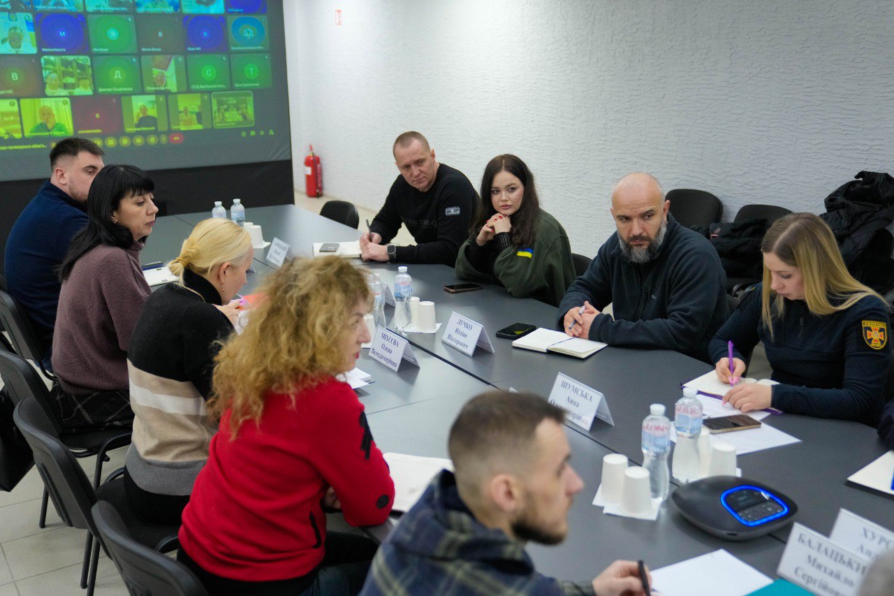 Учасники засідання за столом обговорюють питання, використовуючи систему відеоконференцзв'язку. На великому екрані в залі відображаються вікна з дистанційними учасниками зустрічі.