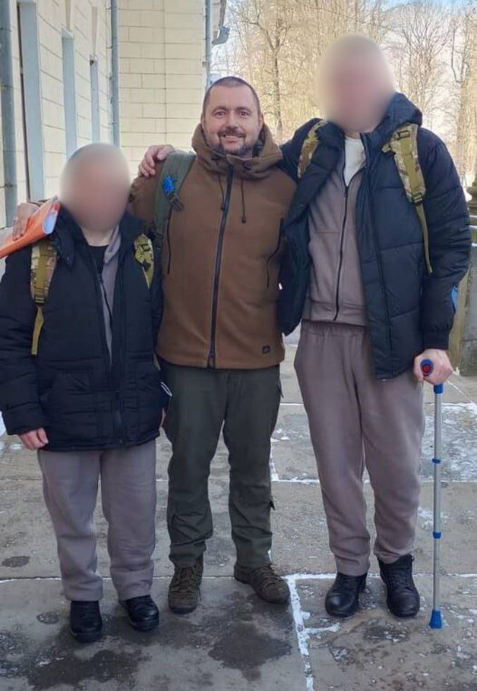 Троє чоловіків стоять поруч на фоні будівлі. Чоловік по центру одягнений у коричневу куртку та штани мілітарі, він обіймає за плечі двох інших супутників. Чоловіки з обох боків мають при собі рюкзаки, а один із них спирається на ліктьову милицю. Обличчя двох осіб на фото приховано.