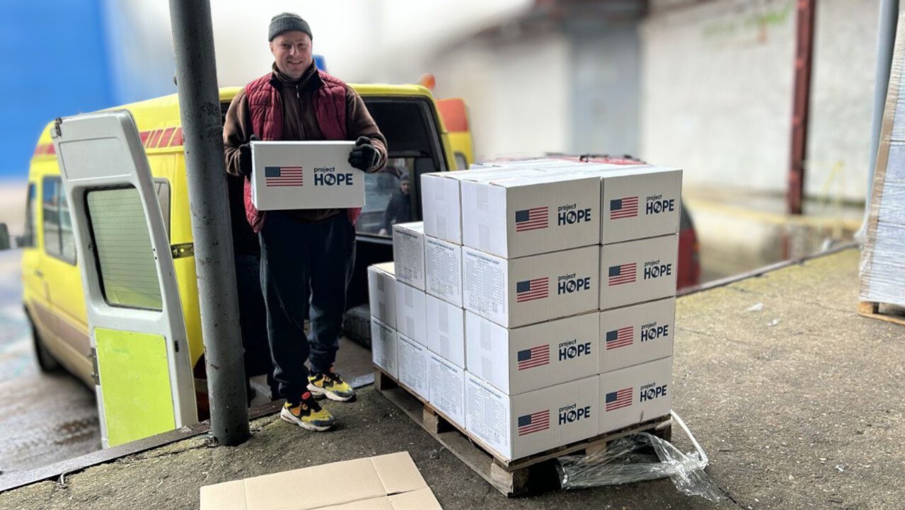Чоловік розвантажує гуманітарну допомогу з жовтого мікроавтобуса. Він тримає в руках білу коробку з логотипом організації «Project HOPE» та прапором США. Поруч на дерев'яному піддоні складена партія аналогічних коробок з гуманітарним вантажем.