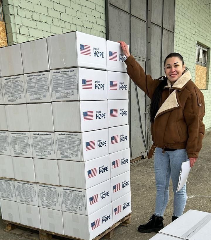 Жінка стоїть поруч із високою палетою гуманітарних вантажів. На білих коробках нанесено логотип «Project HOPE», американський прапор та перелік вмісту гігієнічного набору.