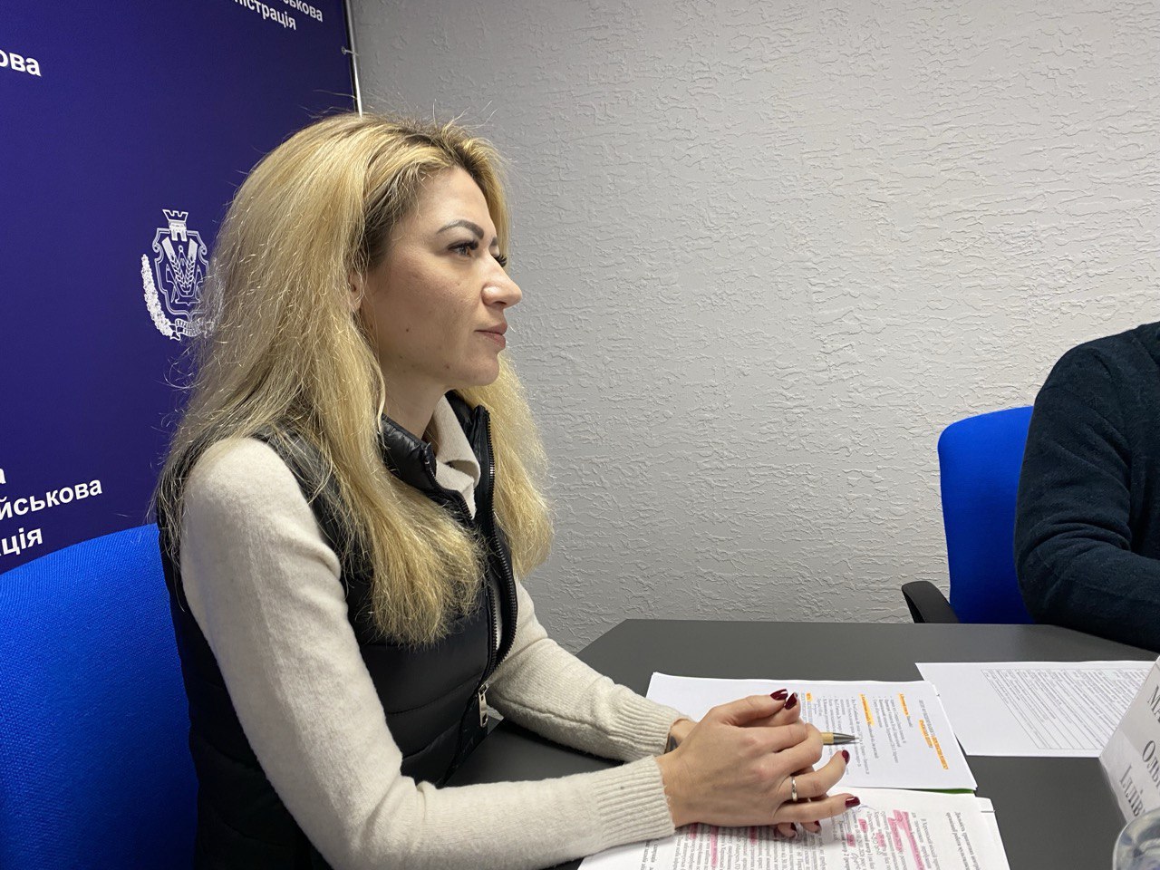 Жінка в офісному приміщенні сидить за робочим столом, на якому лежать документи, на фоні синього брендованого банера військової адміністрації.