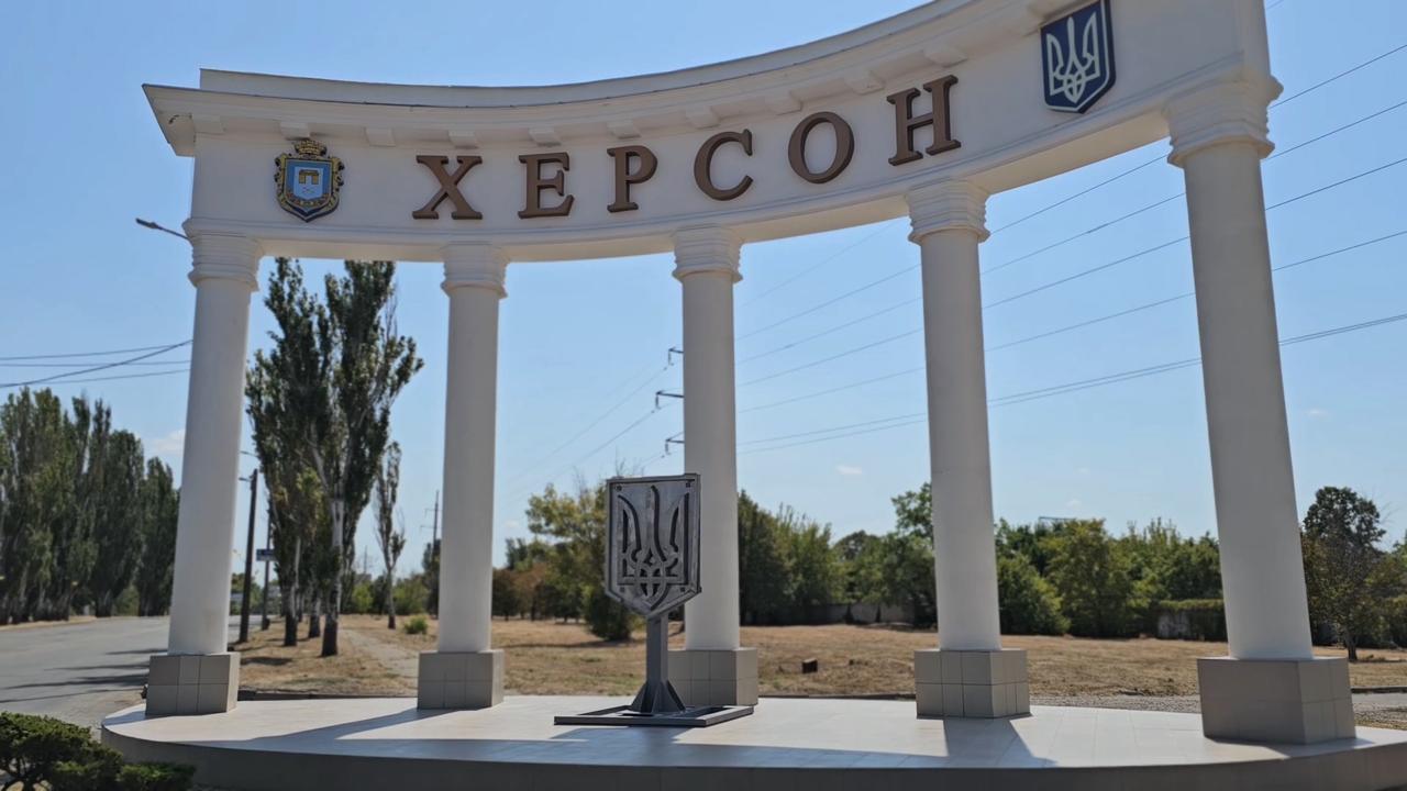 Біла архітектурна споруда у формі півкола з колонами при вході до міста Херсон. На верхній частині розміщено напис назви міста великими літерами, а також герби України та міста. По центру, перед спорудою, встановлено металеву стелу з зображенням Тризуба.