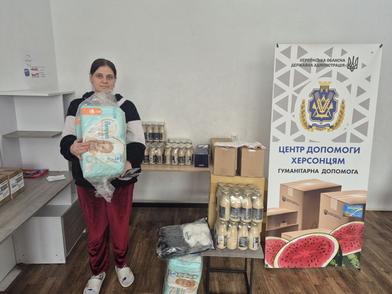 Жінка отримує велику упаковку підгузків у гуманітарному центрі на фоні стелажів із товарами та інформаційного стенда.