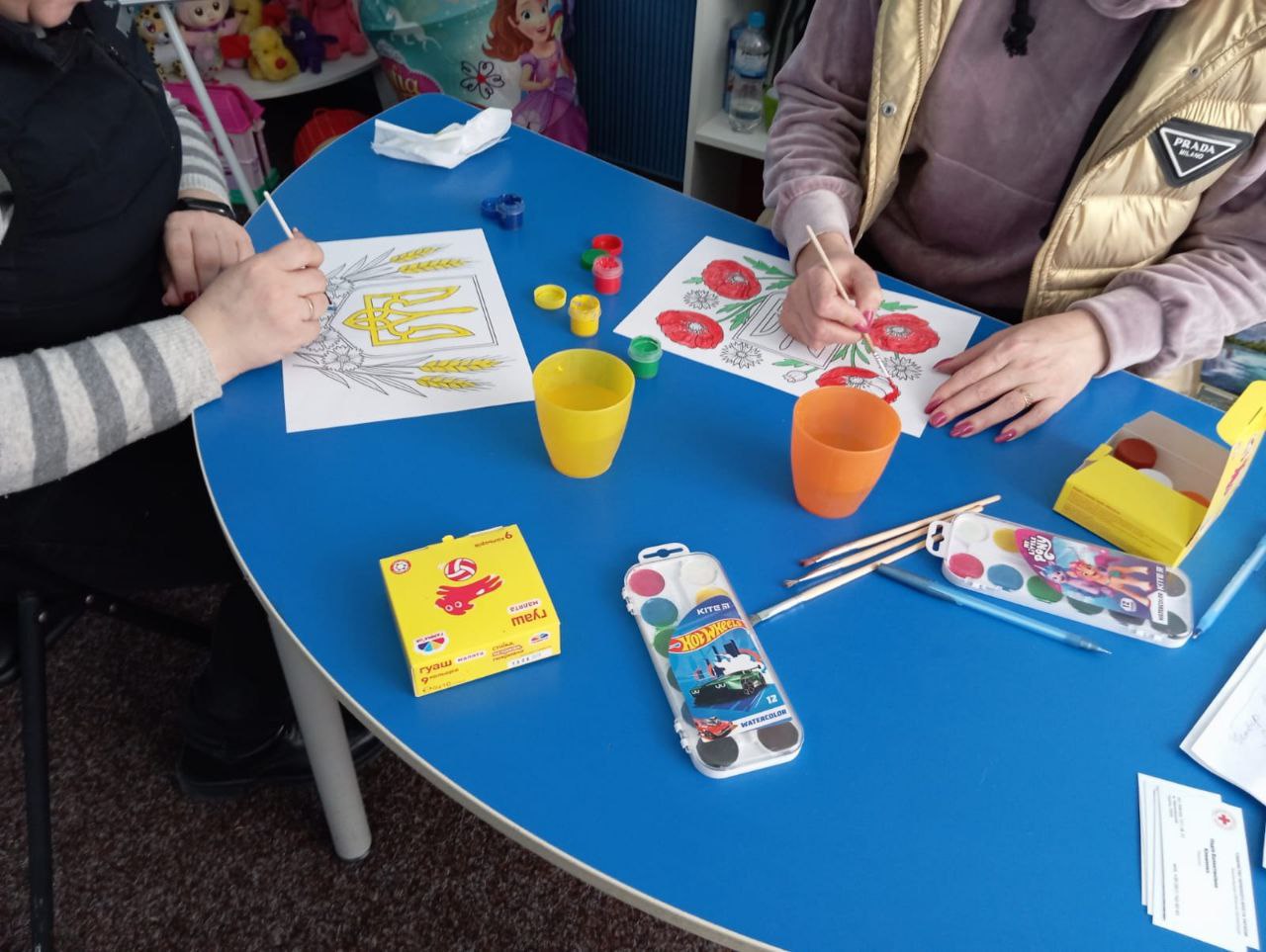 Двоє людей сидять за синім столом і розфарбовують малюнки з національною символікою та квітами маку за допомогою фарб.