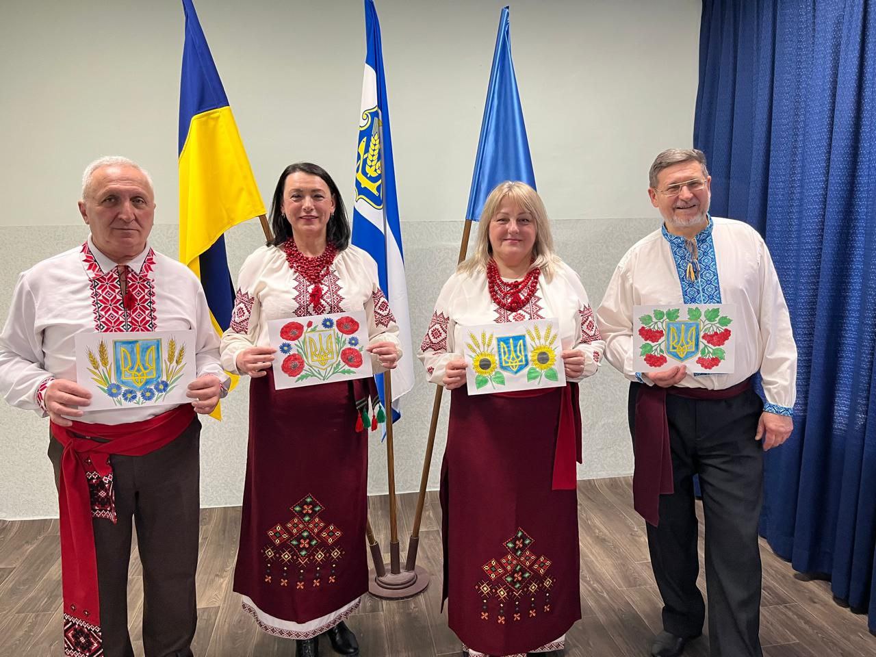 Чотири людини у святкових вишиванках стоять на фоні державних прапорів, тримаючи в руках власноруч розфарбовані малюнки з Гербом України.