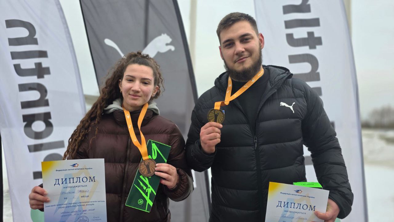 Двоє молодих спортсменів, хлопець та дівчина, позують із нагородами на фоні засніженого поля та брендованих прапорів. Вони тримають у руках дипломи Федерації легкої атлетики України та демонструють медалі на жовтих стрічках. Обоє одягнені в темні зимові спортивні куртки.