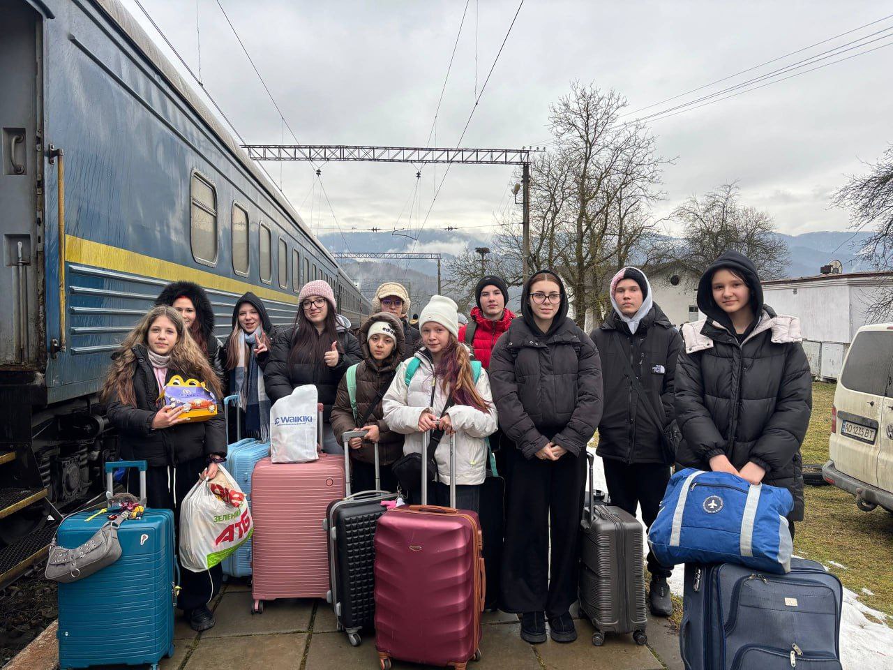 Група дітей та підлітків із валізами й сумками стоїть на пероні поруч із пасажирським потягом. Вони тепло одягнені, деякі тримають пакунки з їжею, на фоні видно залізничні колії та гори в тумані.