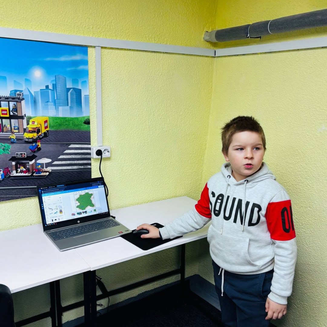 Дитина стоїть біля робочого столу з ноутбуком у навчальному класі. На екрані відкрито графічний інтерфейс програми, поруч на стіні — яскравий плакат із зображенням іграшкового міста.