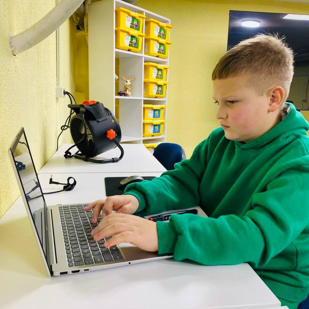 Хлопчик у зеленому худі зосереджено працює за ноутбуком у навчальному класі. Поруч на столі стоїть обігрівач, а на фоні видно білий стелаж із жовтими боксами для речей.