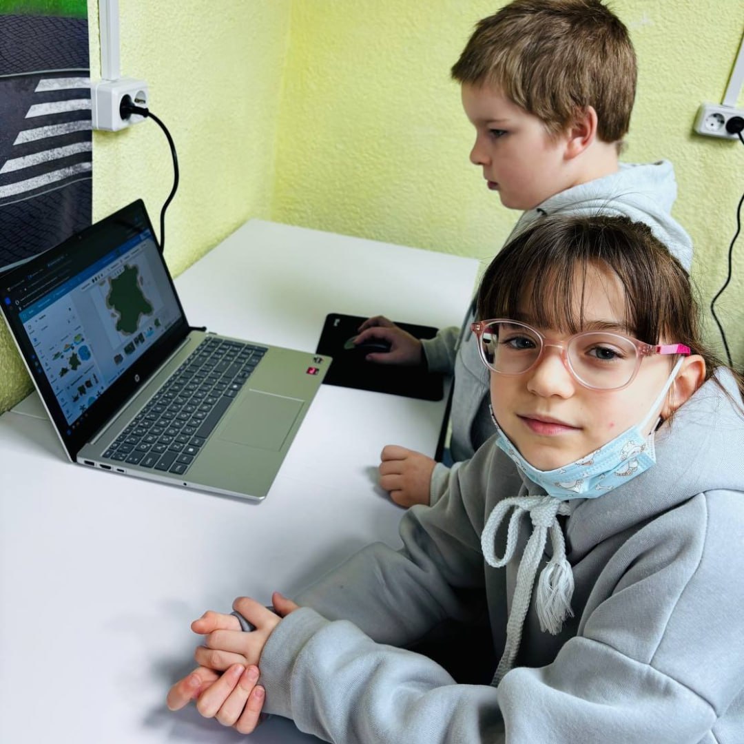 Двоє дітей сидять за робочим столом у навчальному класі. Дівчинка в окулярах та захисній масці дивиться в камеру, а хлопчик поруч працює з ноутбуком. На екрані відкрито графічну програму для моделювання чи гри.