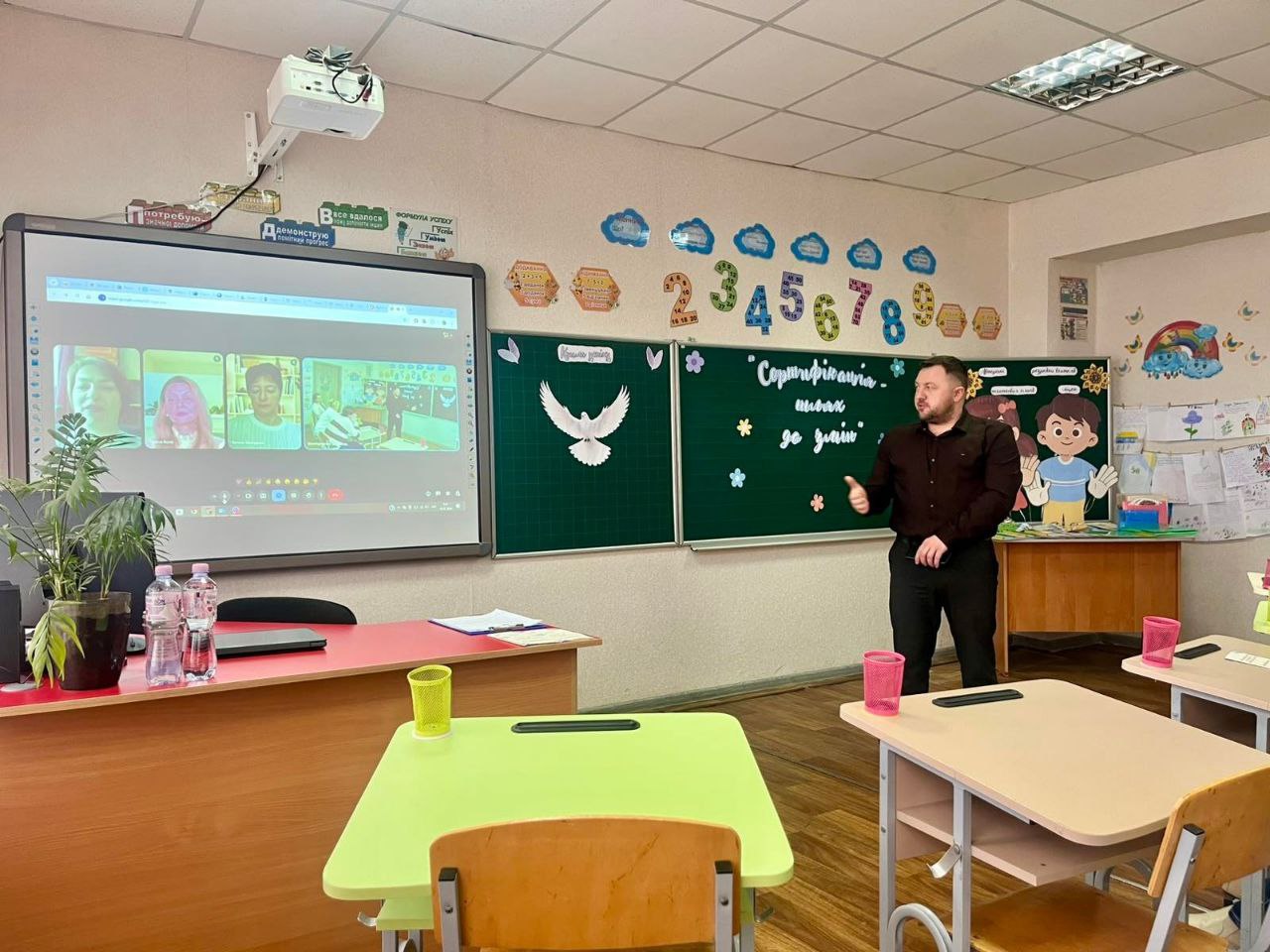Чоловік проводить презентацію в шкільному класі біля інтерактивної дошки, на якій відображено вікно відеоконференції. На задньому плані — зелена шкільна дошка з паперовим голубом та цифрами.