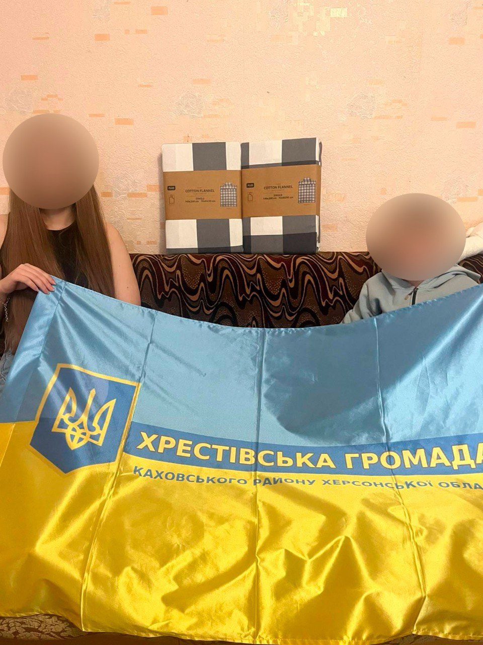 Двоє людей тримають великий прапор Хрестівської громади Каховського району Херсонської області. На фоні, над диваном, стоять два упаковані комплекти постільної білизни. Обличчя людей приховані.