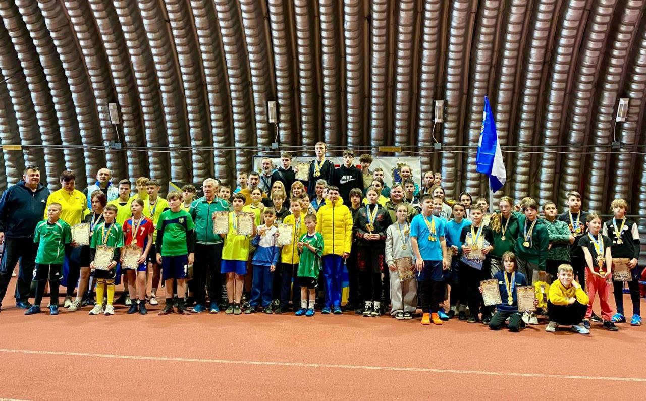 Групове фото учасників спортивних змагань у критому манежі. Велика кількість дітей та підлітків у спортивній формі стоять із медалями та грамотами в руках. Позаду видно державний прапор та елементи конструкції спортивної зали.