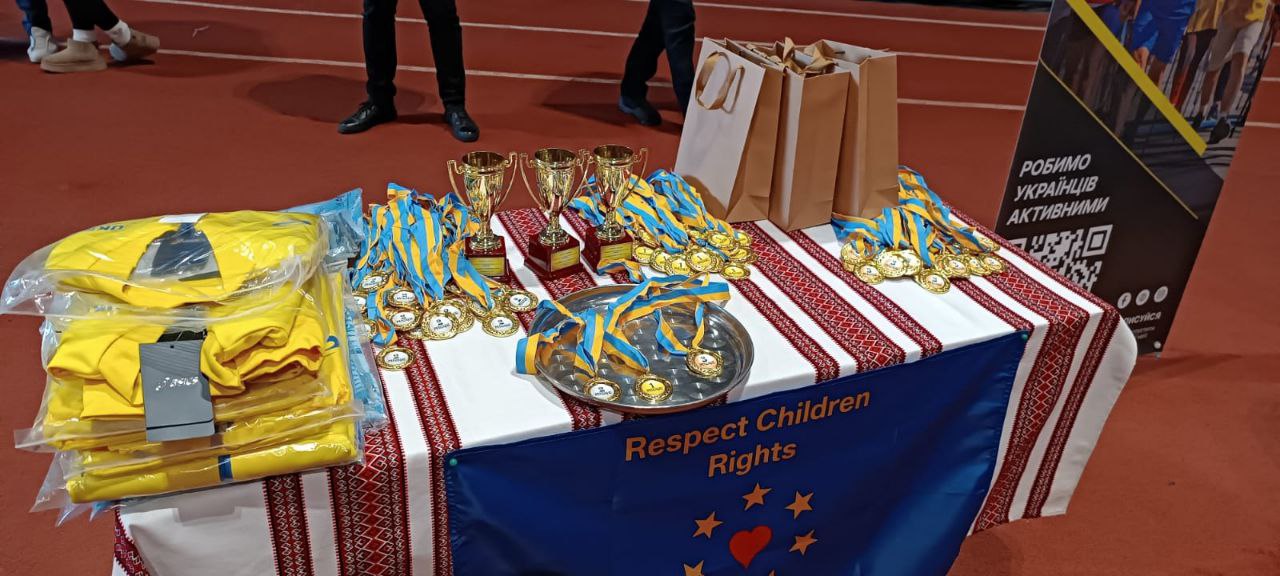 Нагородна атрибутика на столі, застеленому скатертиною з традиційним українським орнаментом. На столі розміщені золоті кубки, медалі на синьо-жовтих стрічках, паперові пакети та стоси складених жовтих футболок. Попереду закріплений синій банер із написом «Respect Children Rights».