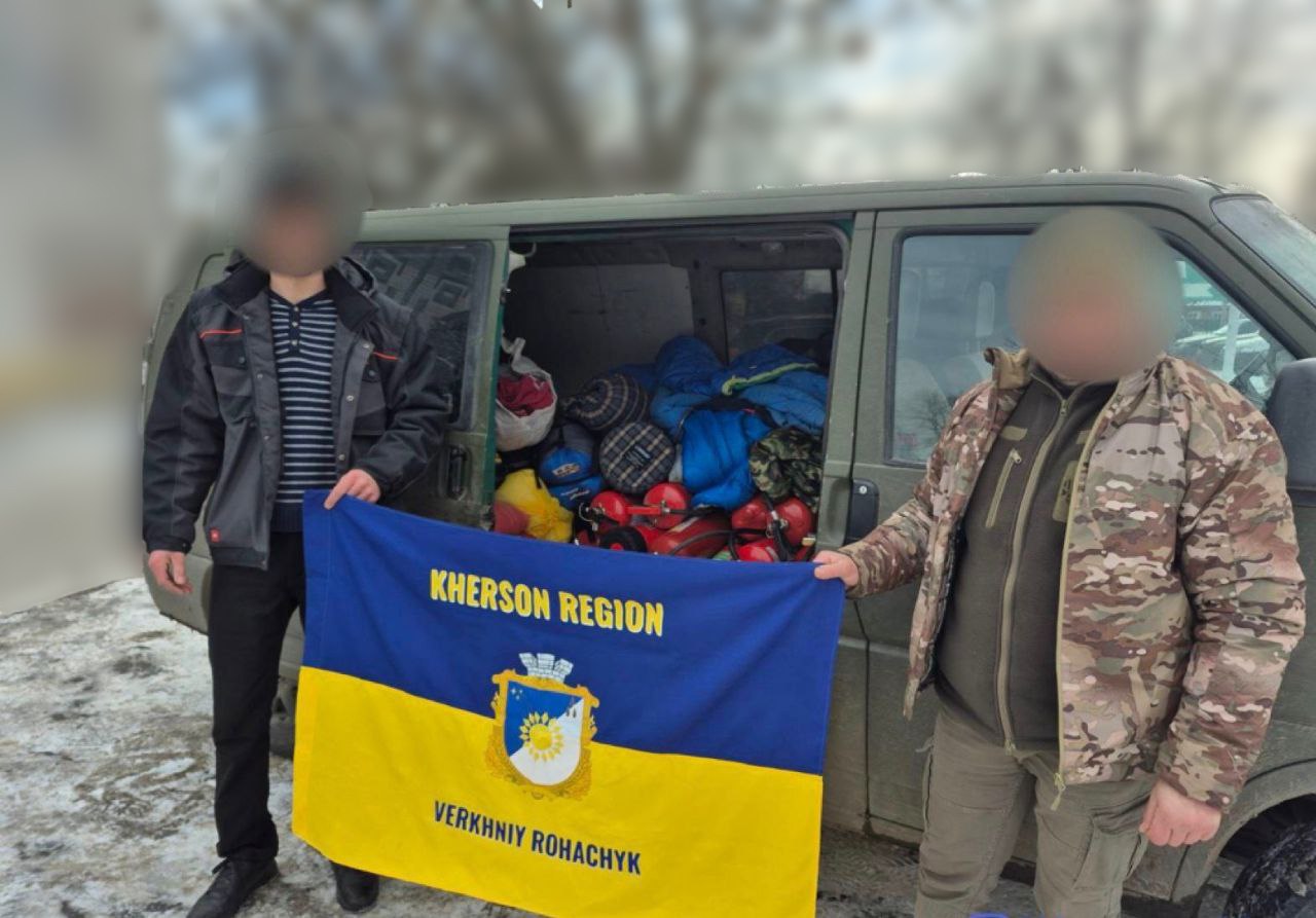 Двоє чоловіків тримають прапор Верхньорогачицької громади (Kherson Region, Verkhniy Rohachyk) на фоні відчиненого мікроавтобуса, завантаженого гуманітарною допомогою: спальними мішками та вогнегасниками. Обличчя людей на зображенні розмиті.