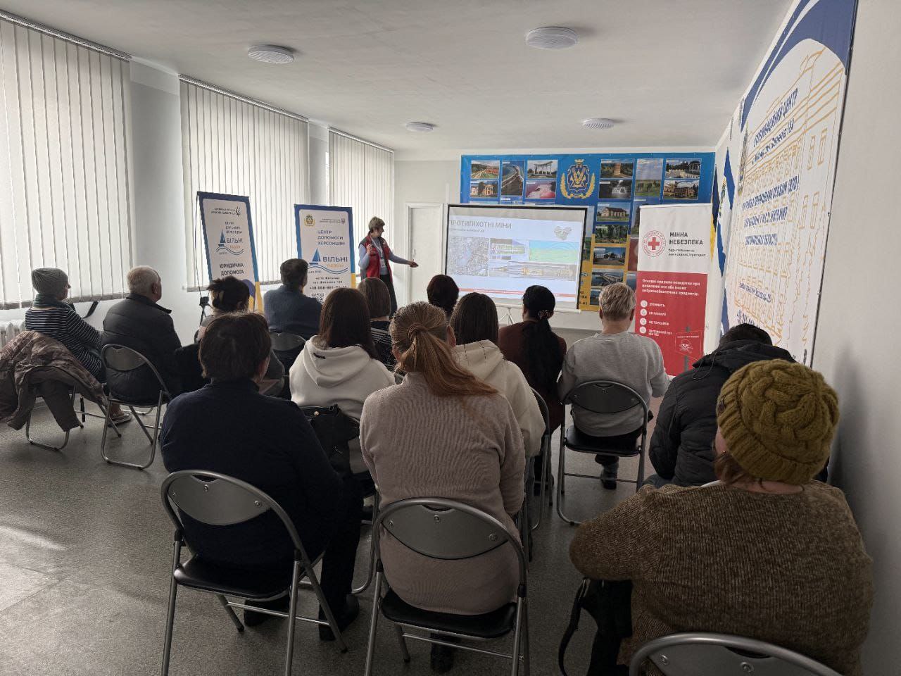 Група людей у світлій залі слухає лекцію про мінну безпеку. Спікерка стоїть біля екрана з презентацією «Протипіхотні міни», поруч розміщено інформаційні банери «Мінна небезпека» та центрів допомоги. Слухачі сидять на стільцях спиною до камери.