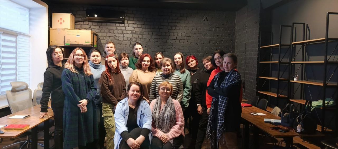 Групове фото людей у сучасному офісному приміщенні з цегляною стіною. Учасники стоять та сидять біля робочих столів, позуючи для спільного знімка в неформальній обстановці.